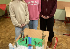 Trzy uczennice prezentują makietę Narnii z rzeką i zimowym krajobrazem wykonanym z naturalnych materiałów.