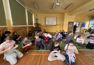 Grupa dzieci siedzi na dywanie w świetlicy i czyta listy, w tle choinka oraz ekran z prezentacją projektu.