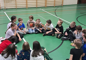 Grupa uczniów siedzi w kręgu na sali gimnastycznej podczas wspólnej zabawy.