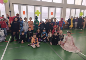 Liczna grupa dzieci w kostiumach karnawałowych siedzi i stoi w sali gimnastycznej podczas wspólnej fotografii.