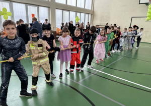Uczniowie w przebraniach stoją w rzędzie i przeciągają linę, a inne dzieci siedzą na ławkach w tle.