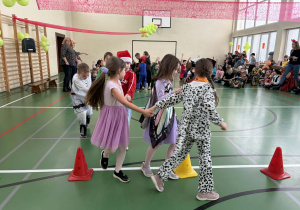 Dzieci w przebraniach trzymają się za ręce i przechodzą między pachołkami podczas zabawy ruchowej w sali gimnastycznej.
