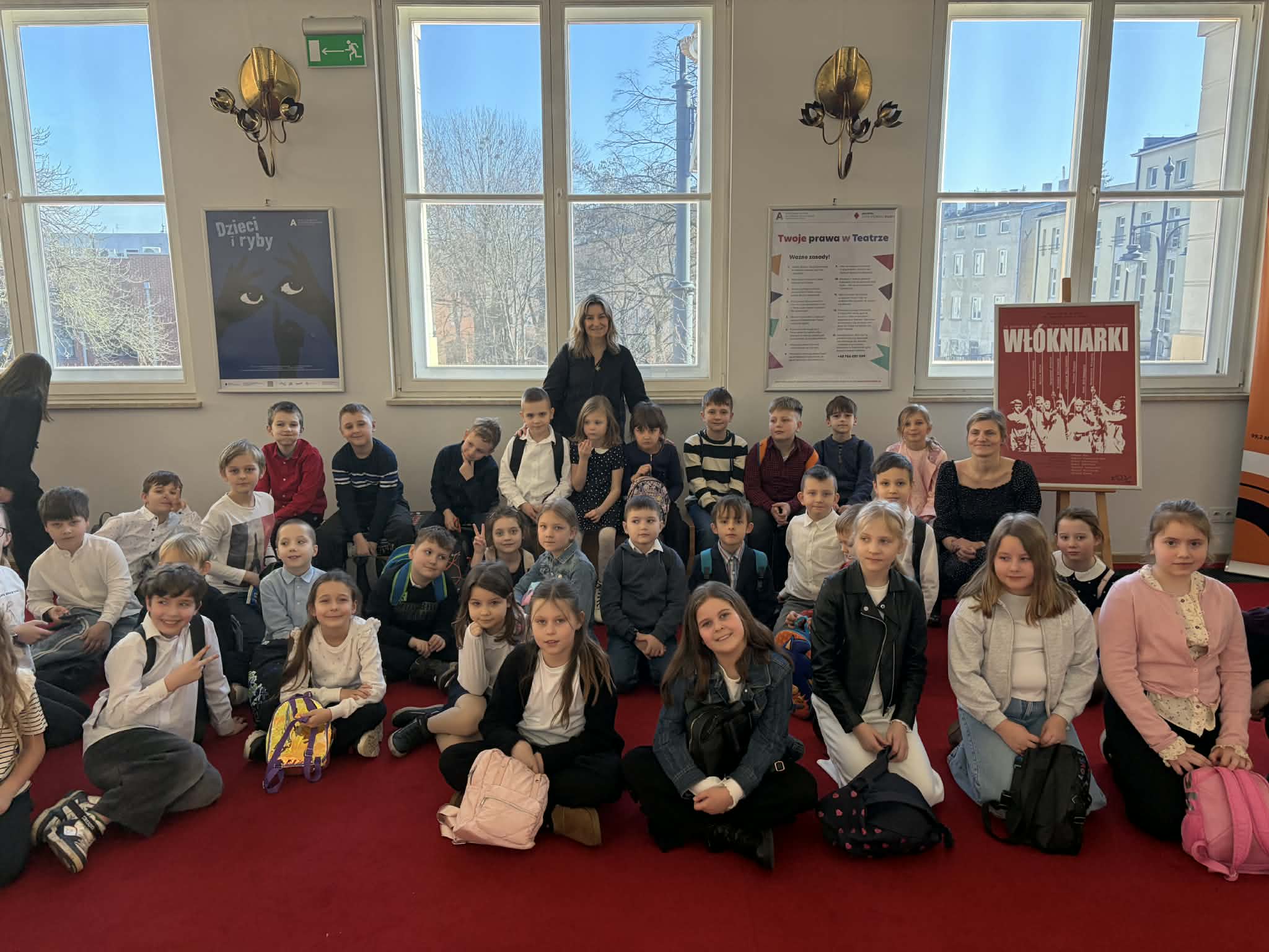 Uczniowie klas 1b i 3b wraz z opiekunkami siedzą na czerwonej wykładzinie w holu Teatr Arlekin po obejrzeniu spektaklu &bdquo;Dzieci i ryby&rdquo;. Dzieci pozują do wsp&oacute;lnego zdjęcia przed dużymi oknami, przez kt&oacute;re wpada jasne światło dzienne. W tle na ścianach widoczne są plakaty teatralne oraz dekoracyjne lampy, a cała grupa uśmiecha się do zdjęcia, tworząc pamiątkę z wizyty w teatrze.