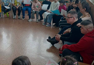 Dzieci i goście siedzą w kręgu w klasie, trzymają rysunki serc i uczestniczą w zajęciach integracyjnych.