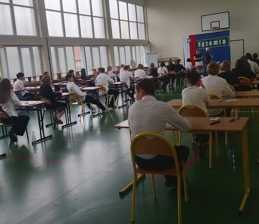 Uczniowie klas ósmych podczas egzaminu z języka polskiego w szkolnej sali gimnastycznej. Na zdjęciu widoczni są uczniowie pracujący w skupieniu nad arkuszami egzaminacyjnymi.