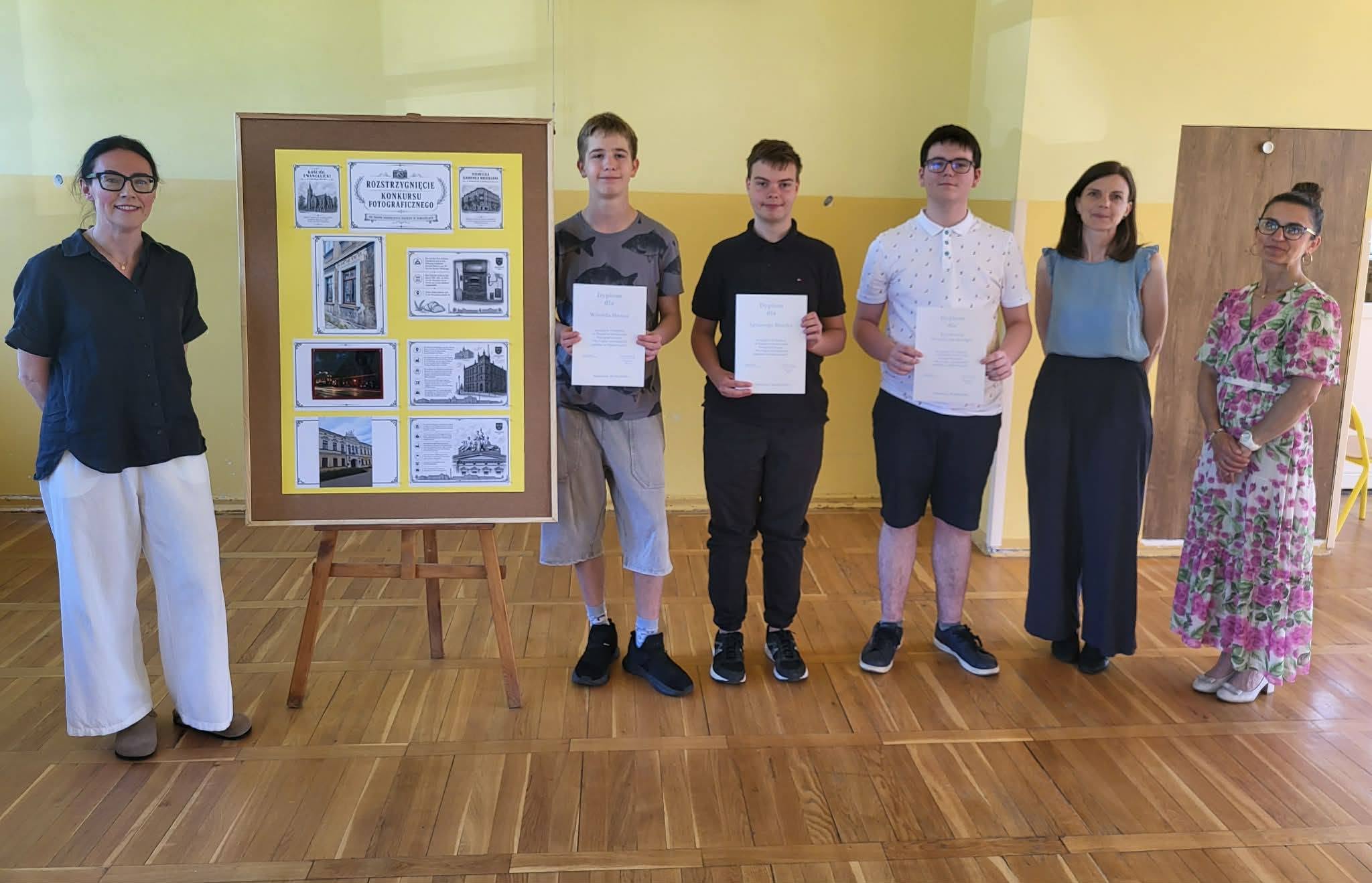 Laureaci miejskiego konkursu fotograficznego wraz z organizatorami stoją obok wystawy nagrodzonych prac i prezentują otrzymane dyplomy.