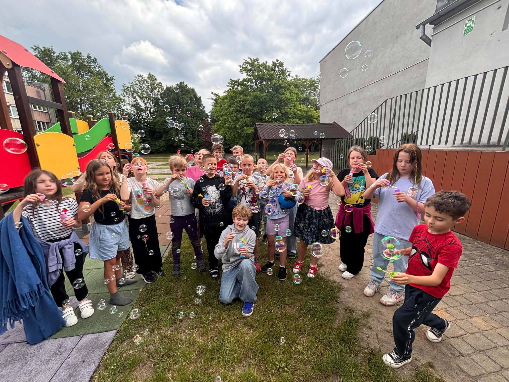 Grupa uczni&oacute;w podczas zabawy z bańkami mydlanymi na szkolnym placu z okazji Dnia Dziecka. Dzieci stoją przed placem zabaw i tworzą kolorowe bańki unoszące się w powietrzu.