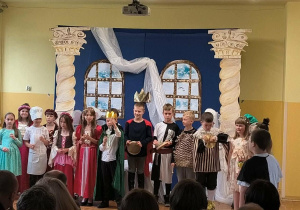 Grupa uczniów klasy 2b w kostiumach teatralnych stoi na scenie po zakończeniu przedstawienia.