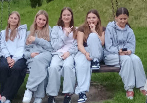 Pięć dziewcząt siedzi obok siebie na dużej drewnianej huśtawce zawieszonej na łańcuchach, umieszczonej na trawiastej skarpie. Cztery z nich mają na sobie luźne, szare dresy, a jedna z dziewcząt (druga z prawej) ubrana jest w ciemny T-shirt i spodnie dresowe. Dziewczęta uśmiechają się, rozmawiają i patrzą w stronę aparatu, jedna z nich trzyma telefon. Scena sprawia wrażenie relaksu i koleżeńskiej atmosfery na świeżym powietrzu.