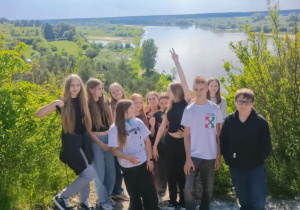 Grupa nastolatków pozuje do wspólnego zdjęcia na tle malowniczego krajobrazu z rzeką i zielonymi łąkami. Uczniowie stoją na skalistym wzniesieniu, wokół nich roślinność i krzewy. Część z nich uśmiecha się, jedna osoba unosi rękę w geście "victory", a pozostali patrzą w stronę aparatu. W tle rozciąga się panoramiczny widok na wodę, pola i lasy pod niebem z częściowo zachmurzonymi chmurami. Zdjęcie ma charakter pamiątkowy z wycieczki lub wyprawy szkolnej.