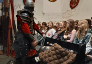 Zdjęcie przedstawia scenę edukacyjną wewnątrz muzeum historycznego. Rycerz w pełnej zbroi i czerwonym stroju prowadzi prezentację dla grupy młodzieży siedzącej w ławkach. Uczniowie uważnie słuchają i uśmiechają się. Na ławce przed nimi leży brązowy, pikowany kaftan ochronny oraz elementy uzbrojenia, w tym hełm. W tle widoczne są średniowieczne tarcze i herby zawieszone na ścianach.