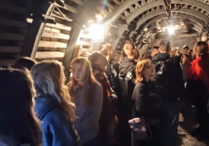 Grupa młodzieży znajduje się wewnątrz podziemnego tunelu, prawdopodobnie w kopalni lub muzeum górnictwa. Tunel jest wzmocniony metalowymi łukami podporowymi i oświetlony lampami zawieszonymi na suficie. Uczniowie ubrani są w ciemne i sportowe ubrania, stoją wzdłuż ścian i słuchają przewodnika. Na podłodze widać wilgoć, a otoczenie ma industrialny, surowy charakter. Zdjęcie oddaje atmosferę zwiedzania miejsca historycznego lub technicznego.