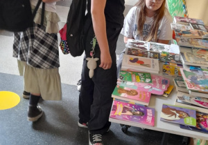 Kilka uczennic ogląda książki i czasopisma rozłożone na stole w szkolnym korytarzu. Jedna z dziewczynek siedzi za stołem, prawdopodobnie obsługując stoisko. W tle widoczne są kolorowe dekoracje, gazetki ścienne i plansze edukacyjne.