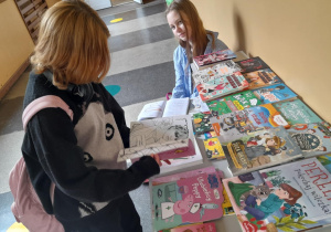Dwie uczennice przy stole z książkami w szkolnym korytarzu. Jedna z dziewczynek siedzi i uśmiecha się, a druga przegląda książkę z czarno-białym rysunkiem. Na stole leży wiele kolorowych książek i publikacji dla dzieci i młodzieży.