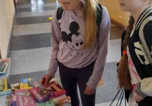 Dwie uczennice stoją przy stole z kolorowymi książkami w szkolnym korytarzu. Jedna z nich przegląda książkę, a druga patrzy na stół. W tle widać jasny korytarz z oknami i tablicami informacyjnymi na ścianie.