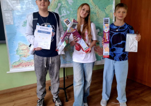Trzech uczniów pozuje do zdjęcia w klasie na tle mapy Europy. Każdy trzyma nagrody: dyplomy, zestawy przyborów szkolnych i edukacyjne gry planszowe. W tle widoczna jest mapa ścienna oraz globusy na szafie.