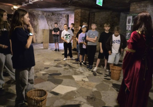 Grupa dzieci zwiedza podziemia z przewodniczką ubraną w historyczny strój. Dzieci stoją w szeregu na kamiennej posadzce w surowym, kamiennym wnętrzu z widoczną ceglaną ścianą i oświetleniem pod sufitem. Przed niektórymi stoją kosze wiklinowe.