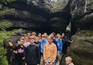 Grupa dzieci stoi na drewnianym pomoście wśród dużych formacji skalnych w otoczeniu lasu. W tle widoczne są drzewa, a niektóre dzieci opierają się o skały. Po prawej stronie na skale siedzi mały chłopiec. To zdjęcie z wycieczki przyrodniczej w terenie górzystym.