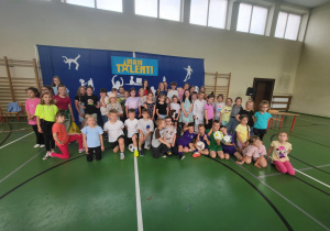 Grupa dzieci pozuje do wspólnego zdjęcia na sali gimnastycznej. W tle znajduje się niebieska dekoracja z napisem ‘Mam Talent!’ oraz białymi sylwetkami przedstawiającymi różne aktywności. Dzieci są ubrane w kolorowe stroje i stoją lub siedzą na zielonej podłodze sportowej.