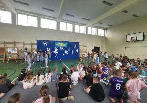 Dzieci zgromadzone na sali gimnastycznej oglądają występ rówieśników na tle niebieskiej dekoracji z napisem ‘Mam Talent!’. Na scenie grupa uczniów w kowbojskich strojach prezentuje swój numer. Po lewej stronie widać sztalugi z pracami plastycznymi, a po prawej uczniowie siedzący na materacach i podłodze.