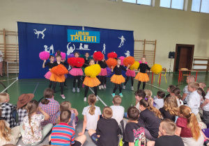 Grupa dziewczynek w kolorowych spódniczkach i z pomponami występuje na scenie podczas szkolnej imprezy ‘Mam Talent!’. Tło sceny jest niebieskie z żółtym napisem ‘Mam Talent!’ i białymi sylwetkami przedstawiającymi różne aktywności. Przed sceną siedzi liczna publiczność złożona z dzieci.