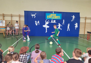 Trzech chłopców prezentuje umiejętności piłkarskie na scenie podczas szkolnego wydarzenia ‘Mam Talent!’. Dwóch ubranych w fioletowe stroje stoi przy piłce, trzeci w zielonym stroju wykonuje drybling. W tle niebieska dekoracja z napisem ‘Mam Talent!’ i białymi sylwetkami symbolizującymi różne pasje. Po lewej stronie siedzą trzy osoby przy sztalugach z rysunkami, a z przodu widoczna jest publiczność dziecięca.