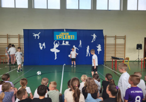 Chłopcy prezentują swoje umiejętności z piłką nożną na scenie szkolnego wydarzenia ‘Mam Talent!’. W tle znajduje się niebieskie tło z napisem ‘Mam Talent!’ i białymi sylwetkami symbolizującymi różne zainteresowania, takie jak taniec, gra na skrzypcach, malowanie czy deskorolka. Przed sceną siedzi publiczność złożona z uczniów.