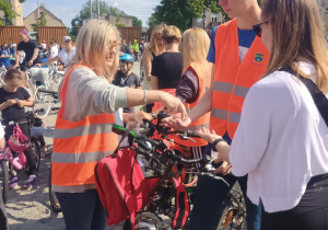 Kilka osób w pomarańczowych kamizelkach odblaskowych rozdaje opaski uczestnikom rajdu rowerowego. W tle widoczni są rowerzyści, dzieci i dorośli, zgromadzeni na placu w słoneczny dzień. Część osób trzyma rowery, inni noszą kaski.