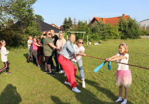 Grupa dorosłych i dzieci bierze udział w przeciąganiu liny na zielonym boisku podczas słonecznego dnia. Po jednej stronie liny stoi dziewczynka w kolorowej tiulowej spódniczce i białej koszulce, uśmiechnięta i trzymająca linę z niebieską wstążką. Po drugiej stronie znajduje się grupa osób – rodzice i dzieci – wspólnie ciągnących linę, uśmiechniętych i zaangażowanych w zabawę. W tle widoczne są drzewa, budynki mieszkalne i bramka do piłki nożnej. Atmosfera jest radosna i integracyjna.