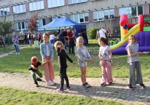 Na zdjęciu grupa dzieci przeciąga linę podczas pikniku szkolnego. Wśród uczestników jest chłopiec w masce Spider-Mana, dziewczynka z pomalowaną twarzą oraz inne dzieci ubrane kolorowo i uśmiechnięte. W tle widać dużą, kolorową dmuchaną zjeżdżalnię, niebieski namiot z napisem „Park Trampolin Air Jump” oraz osoby uczestniczące w wydarzeniu. Z tyłu znajduje się budynek szkolny. Atmosfera jest radosna i pełna dziecięcej energii.