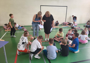 Zdjęcie wykonane w sali gimnastycznej szkoły podstawowej. Na pierwszym planie grupa dzieci siedzi na podłodze w kręgu i rozmawia, obok nich stoją dwie nauczycielki z kartkami w rękach. W tle widać inne dzieci, które w parach lub grupach wykonują zadania plastyczne na dużych arkuszach papieru. Sala ma zieloną podłogę z liniami boiskowymi i jasną ścianę z tablicą do koszykówki.