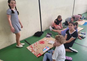 Grupa dzieci pracuje nad plakatami edukacyjnymi wzdłuż ściany sali gimnastycznej. Na pierwszym planie troje uczniów tworzy plakat z hasłem „Udzielanie pierwszej pomocy może uratować życie”, który zawiera czerwony krzyż, serce i rysunki apteczek. Dziewczynka w sukience w paski stoi obok pracy, pozując do zdjęcia. Pozostali uczniowie siedzą lub klęczą, rysując i malując. W tle widać inne grupy dzieci pochylone nad swoimi projektami.
