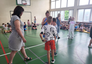 Sala gimnastyczna pełna uczniów. Na pierwszym planie dwóch chłopców stoi naprzeciw nauczycielki w niebieskiej sukience, która wręcza im nagrody. Obok stoi druga nauczycielka w jasnej sukience. W tle dzieci siedzą na podłodze, obserwując wydarzenie. Niektóre osoby rozmawiają, inne czekają w kolejce. W sali widać kosz do koszykówki i duże okna wpuszczające światło dzienne.