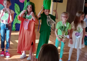 Sześć dziewczynek stoi na scenie w przebraniach symbolizujących warzywa. Każda z nich ma na ubraniu przypiętą ilustrację z uśmiechniętym warzywem, np. pomidorem, marchewką, pietruszką, szczypiorkiem i burakiem. Dzieci mają kolorowe stroje – w odcieniach zieleni, czerwieni, pomarańczu i bieli – oraz elementy dekoracyjne, np. peruki z liści czy korony z trawy. W tle widoczne są dekoracyjne kwiaty i ciemnoniebieska kotara.