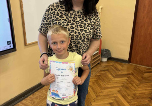 Zdjęcie przedstawia ucznia Jakuba Bednarka stojącego obok nauczyciela bibliotekarza, pani Moniki Juran-Tłoczek, w klasie szkolnej. Jakub trzyma w rękach kolorowy dyplom i uśmiecha się. Ma na sobie jasnożółtą koszulkę i krótkie spodenki z nadrukiem nosorożców. Pani Monika, ubrana w bluzkę w panterkę i dżinsy, obejmuje go delikatnie ramieniem i również się uśmiecha. W tle widoczne są: tablica, mapa Europy, drewniana podłoga i fragment ekranu multimedialnego.