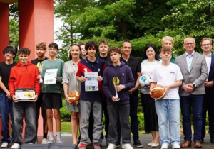 Grupowe zdjęcie przedstawiające uczniów i nauczycieli pozujących z pucharami, piłkami i dyplomami na tle czerwonego budynku i zielonych drzew. Uroczystość odbyła się 24 czerwca w ogrodzie Młodzieżowego Domu Kultury w Pabianicach z okazji rozdania nagród za współzawodnictwo sportowe szkół Powiatu Pabianickiego w roku szkolnym 2024/2025. Osoby na zdjęciu są uśmiechnięte, a część z nich trzyma trofea sportowe. Wśród obecnych znajdują się nauczyciele wychowania fizycznego – Sebastian Grzelczyk i Rafał Raźniewski.