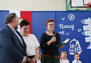 Na zdjęciu troje przedstawicieli Rady Rodziców. Kobieta w ciemnym swetrze i okularach przemawia do mikrofonu, trzymając otwarty zeszyt. Obok niej stoją kobieta w białej bluzce oraz mężczyzna w garniturze w prążki. W tle widoczna dekoracja z napisem „Ruszaj w swoją podróż” oraz uczniowie siedzący na podłodze.