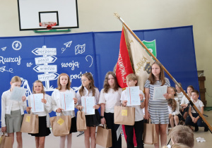 Zdjęcie przedstawia grupę wyróżnionych uczniów klasy 4b stojących na tle niebieskiej dekoracji z hasłem „Ruszaj w swoją podróż” i drogowskazem z napisem: marzenia, pasja, wiedza, przygoda. Uczniowie ubrani są w galowe stroje – białe koszule i bluzki, ciemne spódniczki lub spodnie – i trzymają w rękach świadectwa oraz papierowe torby z nagrodami. W tle widoczny jest poczet sztandarowy z czerwono-kremowym sztandarem szkoły oraz herbem Szkoły Podstawowej nr 13 w Pabianicach. W prawym dolnym rogu częściowo widoczna jest grupa siedzących uczniów.