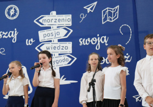 Zdjęcie przedstawia sześcioro uczniów klas trzecich występujących na scenie podczas uroczystości zakończenia roku szkolnego w Szkole Podstawowej nr 13 w Pabianicach. Troje dzieci trzyma mikrofony i śpiewa, pozostali recytują lub oczekują na swoją kolej. Dzieci ubrane są odświętnie – w białe koszule i ciemne spódnice lub spodnie. W tle widoczna jest niebieska dekoracja z białymi napisami „Ruszaj w swoją podróż” oraz drogowskazem z hasłami: marzenia, pasja, wiedza, przygoda. Dekorację uzupełniają rysunki papierowych samolotów i mapy.