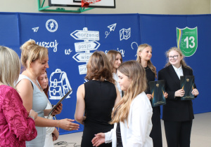 Zdjęcie przedstawia moment wręczania nagród uczniom klasy 8a. Po lewej stronie kadru dyrektor szkoły, pani Anna Michalak, ubrana w jasnoniebieską sukienkę, ściska dłoń jednej z uczennic. Obok niej stoi wicedyrektor Edyta Makowska w różowej sukience z koronkowym wzorem. Po prawej stronie widać grupę wyróżnionych uczennic w eleganckich strojach – białych koszulach, czarnych sukienkach i garniturach – trzymających czarne teczki z napisem „Świadectwo ukończenia szkoły podstawowej” oraz godłem Polski. W tle widoczna jest dekoracja z niebieskim tłem, białym drogowskazem i hasłem „Ruszaj w swoją podróż” oraz herbem szkoły.