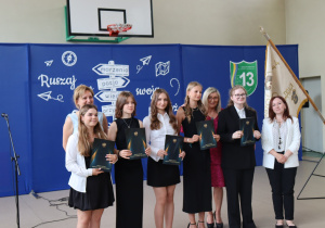Zdjęcie przedstawia grupę wyróżnionych uczennic klasy 8a pozujących do pamiątkowego zdjęcia po wręczeniu świadectw ukończenia szkoły podstawowej. Uczennice ubrane są elegancko w czarne lub białe stroje i trzymają teczki z godłem Polski i napisem „Świadectwo ukończenia szkoły podstawowej”. Po lewej stronie stoi dyrektor szkoły, pani Anna Michalak, w jasnoniebieskiej sukience. Obok niej znajdują się wicedyrektor Edyta Makowska w bordowej sukience oraz wychowawczyni klasy Magdaleną Maciszewską w białej marynarce. W tle widoczna jest dekoracja z hasłem „Ruszaj w swoją podróż” oraz herbem szkoły i pocztem sztandarowym. Atmosfera jest uroczysta i pełna dumy.