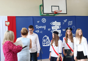 Zdjęcie przedstawia moment wręczenia nagród wyróżnionym uczniom klasy 8b. Po lewej stronie kadru widoczne są plecy dwóch kobiet – dyrektor szkoły Anny Michalak w jasnoniebieskiej sukience i wicedyrektor Edyty Makowskiej w różowej sukience z koronkowym wzorem. Na środku stoi uczeń w białej koszuli i okularach, odbierający gratulacje. Obok niego trzy uczennice – dwie ubrane w galowy strój pocztu sztandarowego z granatowymi beretami i biało-czerwonymi szarfami, a jedna w białej koszuli i czarnej spódnicy. W tle znajduje się dekoracja z drogowskazem i hasłem „Ruszaj w swoją podróż” oraz herbem szkoły. Atmosfera jest uroczysta.