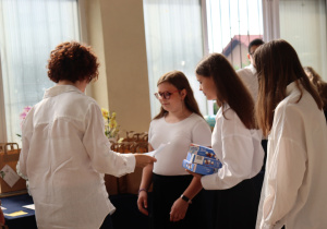Na zdjęciu nauczycielka w białej koszuli, Sylwia Muszyńska, wręcza świadectwa uczennicom klasy 7a. Dziewczęta ubrane są w białe bluzki i czarne spódnice. Jedna z nich trzyma dwa niebieskie pudełka z upominkami. W tle widać stół z torbami prezentowymi oraz ozdobne storczyki.
