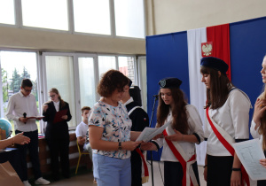 Grupa uczennic w białych bluzkach i czarnych spódnicach odbiera nagrody od nauczycielki w jasnej bluzce we wzory i niebieskich spodniach. W tle dekoracja z hasłem „Ruszaj w swoją podróż”, godło Polski oraz sztandar szkoły z napisem „Ojczyzna Nauka Praca”. Po lewej stronie stoją członkowie pocztu sztandarowego w mundurkach szkolnych z czerwonymi szarfami.