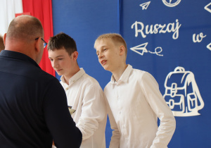 Grupa uczniów w eleganckich koszulach odbiera dyplomy od nauczyciela wychowania fizycznego, ubranego w granatową bluzę. Uczniowie stoją w szeregu, trzymając w rękach dyplomy. W tle widoczna dekoracja z hasłem „Ruszaj w swoją podróż” oraz biało-czerwona flaga z godłem Polski. Uroczystość odbywa się w jasnej sali gimnastycznej z dużymi oknami.