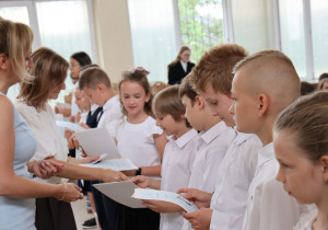 Nauczycielka w jasnoniebieskiej sukience ściska dłoń uśmiechniętej dziewczynki w białej bluzce. Obok stoją uczniowie w galowych strojach, trzymając świadectwa. W tle widać inne dzieci, nauczycieli oraz stoliki z dokumentami i kwiatami.
