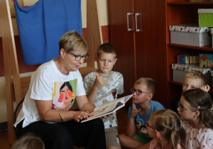 Nauczycielka Monika Metller siedzi w kręgu dzieci i czyta im książkę. Uczniowie uważnie słuchają, siedząc na podłodze w klasie. W tle znajdują się regały z materiałami dydaktycznymi i pudełkami.