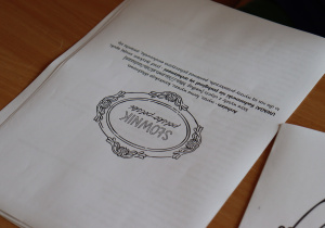 Zbliżenie na kartę pracy zatytułowaną „Słownik podręczny”. Na arkuszu widoczne są ozdobne ramki oraz instrukcja w języku polskim, w której uczniowie proszeni są o wpisanie trudnych słów wraz z definicjami. Kartka leży na drewnianym blacie szkolnej ławki. Widoczna część tekstu zawiera m.in. hasła: „Adrian – wyraz, którym wyrażasz...”, „UWAGA! Rekomendujemy nie posługiwać się przekleństwami...”.