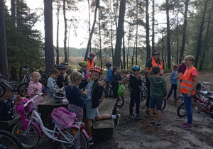 Klasa 2b na przerwie w lesie podczas wycieczki rowerowej. Dzieci jedzą śniadanie i odpoczywają. W okularach i kamizelce odblaskowej wychowawczyni p. Monika Mettler.