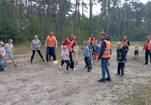 Dzieci z klasy 2b grają w piłkę z wychowawczynią i opiekunami na leśnej polanie. Wszyscy uczestnicy noszą odblaskowe kamizelki i świetnie się bawią.